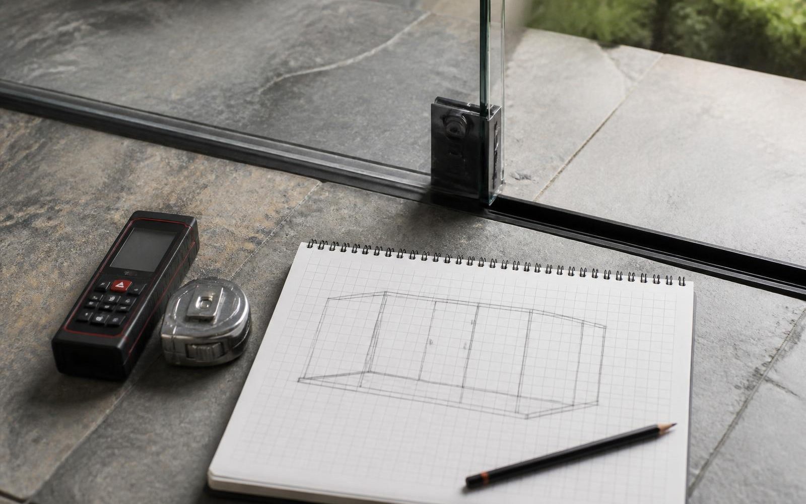 Laser measure, tape measure and a sketched enclosure layout on a patio beside a frameless glass panel and its floor track